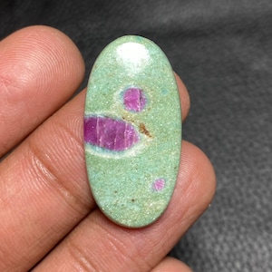 Op de afbeelding: Een ovaalvormige cabochon edelsteen met een gladde, lichtgroene basis en een gespikkelde textuur. De steen bevat drie opvallende, levendige magenta-kleurige insluitingen van verschillende groottes in het groene oppervlak.