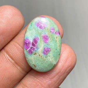 Puede incluir: Gema ovalada con una base verde claro e inclusiones rosas vibrantes. La piedra pulida muestra un patrón natural, con grupos rosas dispersos por la superficie verde. La gema se sostiene en una mano, resaltando su forma y variaciones de color.