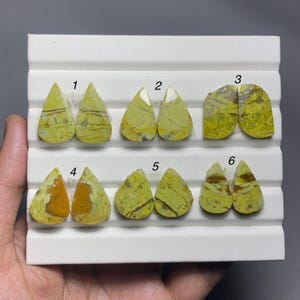 May include: Six pairs of polished, teardrop-shaped yellow and brown patterned cabochons arranged on a white display stand. Each pair is numbered from one to six, showcasing natural stone textures with varied veining and earthy colour tones.