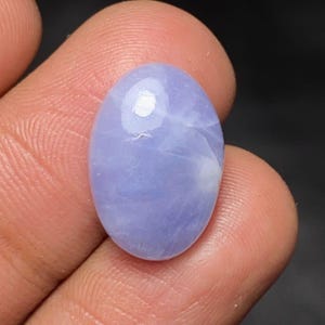 May include: An oval-shaped, smooth, polished gemstone with a light lavender-blue colour. The stone features a translucent appearance with subtle, cloudy white internal inclusions and a reflective surface that catches the light.