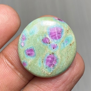 Puede incluir: Una gema redonda con una base de color verde claro, con parches irregulares de azul claro y rosa vibrante. Las áreas rosas tienen una apariencia cristalina, creando un patrón único. La piedra se sostiene en una mano.