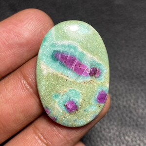 May include: An oval-shaped cabochon gemstone featuring a smooth, polished surface with a light green and mint-colored matrix. The stone is embedded with several distinct, deep magenta-colored ruby inclusions of varying sizes and irregular shapes.