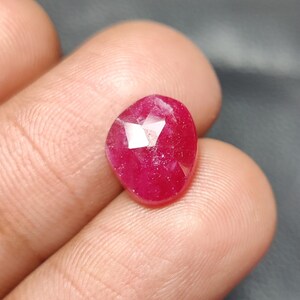 May include: A deep red ruby gemstone with a faceted cut. The gemstone is set in a bezel setting and has a polished finish.
