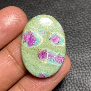 May include: An oval-shaped cabochon gemstone featuring a smooth, light green base color with distinct, irregular inclusions of vibrant magenta-pink mineral deposits scattered across the surface, creating a natural, mottled, and polished appearance.