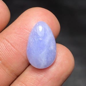May include: A smooth, teardrop-shaped blue lace agate gemstone with a polished surface. The stone features a soft, pale periwinkle blue color with subtle, cloudy white banding patterns visible throughout its translucent, rounded, and symmetrical form.