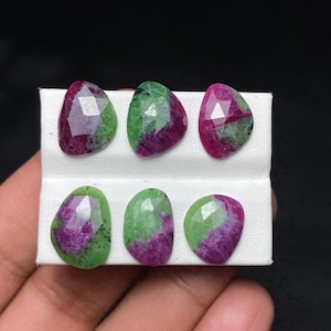 May include: Six faceted gemstones with a mix of deep red and green hues. The stones are oval and teardrop shaped, arranged on a white surface. The gems have a polished, reflective surface.