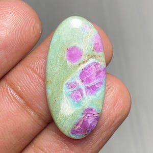 Puede incluir: Una gema ovalada con una base verde claro e inclusiones rosas y moradas vibrantes. La piedra pulida se sostiene en una mano, mostrando su belleza natural. La combinación de colores única de la gema la convierte en una pieza llamativa. Aproximadamente 3 cm de largo.