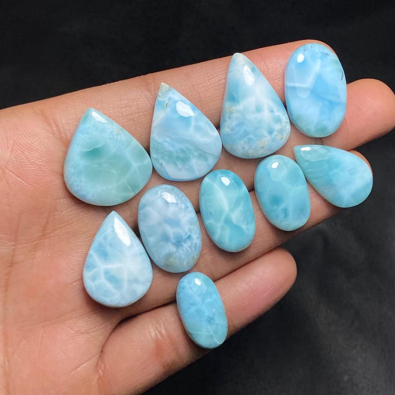 May include: A collection of polished larimar gemstones in various shapes, including teardrops and ovals. The stones display a range of light blue hues with white marbling, resembling ocean waves. The gemstones are held in a hand against a dark background.