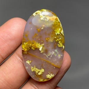 May include: An oval-shaped cabochon gemstone featuring a translucent, pale lavender base colour. The stone is decorated with vibrant yellow and golden-brown moss-like inclusions that create a natural, organic pattern across the smooth surface.