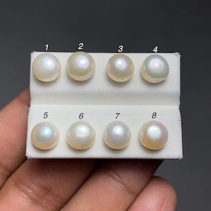 Könnte beinhalten: Acht runde Perlen in verschiedenen Farbtönen werden auf einer weißen Karte präsentiert. Die Perlen reichen von cremeweiß bis irisierend, mit subtilen rosa und grünen Farbtönen. Die Perlen sind von 1 bis 8 nummeriert.