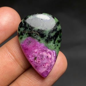 May include: A teardrop-shaped polished gemstone featuring a vibrant magenta-pink section contrasted with a deep forest green and black speckled area. The surface is smooth and reflective, showcasing natural mineral inclusions and colour variations.
