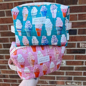 May include: Two quilted cosmetic bags, one teal and one pink, are stacked. Both bags feature a repeating pattern of ice cream cones in various colors. Each bag has a small handle and a label that reads "Handmade with love, Melanie Covington."