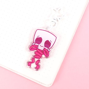 May include: A clear acrylic keyring featuring a cartoon character with a white head and pink arms and legs. The character is holding its face with its hands. The keyring is on a white notebook with a pink background.