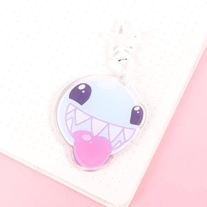May include: A clear acrylic charm with a cartoon monster face. The charm has a light blue body, purple eyes, and a pink tongue. It is attached to a white notebook on a pink background.