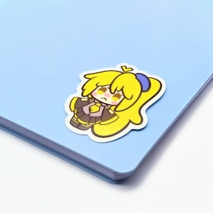May include: A die-cut sticker featuring a cartoon character with yellow hair, a blue hair tie, and a brown and yellow outfit. The sticker is placed on a light blue notebook. The character has a slightly sad expression.