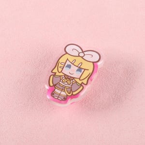 May include: A cartoon character pin with a white bow, blonde hair, and a pink border. The character is wearing a detailed outfit with shades of grey, gold, and pink. The pin is set against a soft pink background.