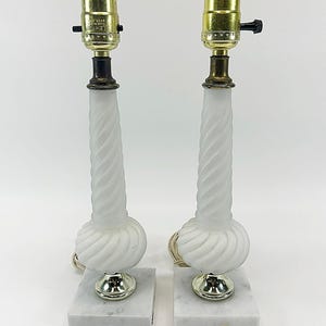 May include: A pair of table lamps featuring frosted white glass bodies with a twisted, spiral design. Each lamp sits on a square white marble base with grey veining and includes a brass-colored metal socket with a black turn-knob switch.