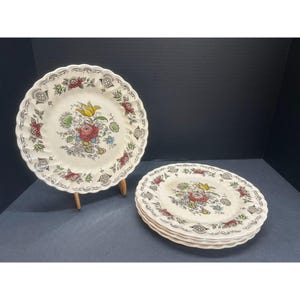 May include: Set of vintage floral pattern plates. The plates have a cream-colored background with a scalloped edge. The floral design includes red, yellow, and blue flowers. One plate is displayed on a wooden stand, while the others are stacked.