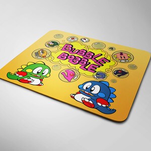 Può includere: Tappetino per mouse giallo con un personaggio dei cartoni animati che soffia una bolla e il testo "Bubble Bobble".