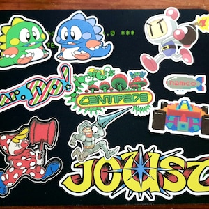 May include: A collection of colourful vintage arcade game stickers featuring characters like Mr. Do!, Centipede, Joust, and a Pac-Man-like character. The stickers are on a brown surface.