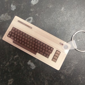 Puede incluir: Un llavero beige y marrón Commodore 64 con un anillo. El llavero tiene la forma de una computadora vintage con un teclado marrón y una carcasa beige. El texto "Commodore 64" está impreso en la carcasa.