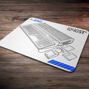 Puede incluir: Un ratón de ordenador blanco con una ilustración en blanco y negro de un ordenador Atari 1040ST, un teclado, un ratón y disquetes. El texto "ATARI" y "1040ST" está impreso en el ratón de ordenador.