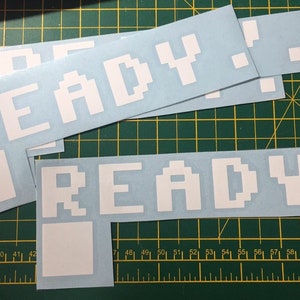 May include: Multiple white vinyl stickers, each displaying the word "READY" in a pixelated font. Each sticker features a square graphic and a dot. The stickers are on a light blue background.