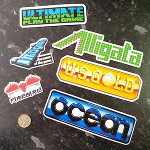 May include: A collection of vintage video game stickers featuring logos for 'Ultimate Play The Game', 'Firebird', 'Ocean', 'US Gold', and 'Alligator'. The stickers are printed on white paper with colourful graphics and text.