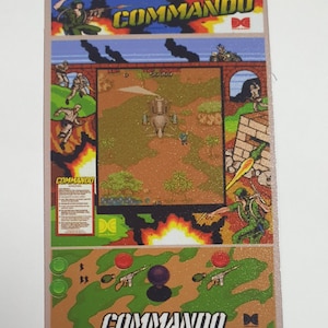 May include: A vintage arcade game control panel featuring a Commando game screen with a helicopter flying over a desert landscape. The panel has a green camouflage background with red and green buttons and a joystick. The text "COMMANDO" is printed in white on the panel.