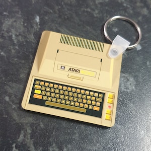 Puede incluir: Un llavero beige y dorado con forma de ordenador Atari 800. El llavero tiene un teclado negro y amarillo con el logotipo de Atari y el texto "ATARI 800" impreso en la parte delantera.