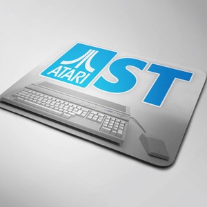 Puede incluir: Una alfombrilla de ratón rectangular con un diseño de ordenador Atari ST. La alfombrilla tiene un fondo gris claro con el logotipo de Atari y "ST" en azul. Un ordenador y un ratón grises están representados en la alfombrilla.