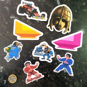 May include: A collection of colourful stickers featuring a red race car, a gold helmet, a pink and orange triangle, a blue character with the text "Monty Mole", a cartoon fox with the text "Sam Stoat Safebreaker", a red karate character, and a blue karate character.