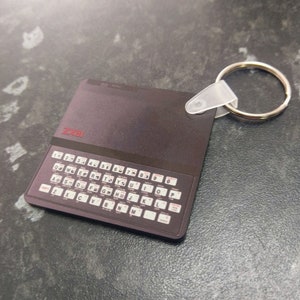 May include: A black and silver keychain shaped like a vintage ZX81 computer with a red keyboard and the text "ZX81" printed on the top.