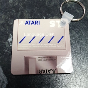 May include: A light pink Atari Language Disk keyring with a silver key ring. The keyring features the Atari logo and the text "Language Disk". The text "MICRO FLOPPY DISK" is also visible.