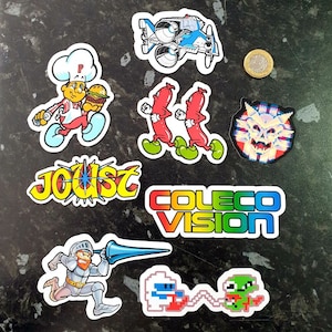 May include: A set of nine colourful stickers featuring retro video game characters and logos. The stickers include a character holding a burger, a spaceship, two hot dogs, a knight, a ghost, a logo for Joust, and a logo for ColecoVision.