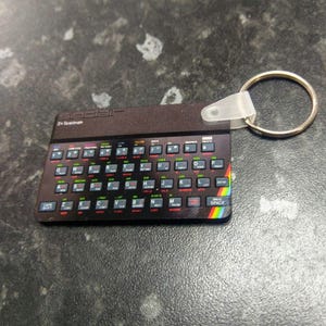 May include: A black and silver keychain featuring a replica of a vintage Sinclair ZX Spectrum computer keyboard. The keychain has a rainbow stripe on the bottom edge.
