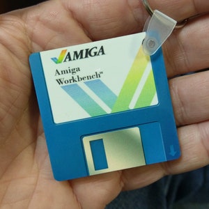 Könnte beinhalten: Ein blauer und weißer Amiga Workbench Floppy Disk Schlüsselanhänger. Der Schlüsselanhänger zeigt das Amiga Logo und den Text "Amiga Workbench".