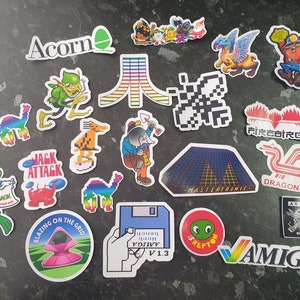 May include: A collection of colourful stickers featuring retro video game and computer logos, including Acorn, Jack Attack, Firebird, Dragon 32, Mastertronic, Amiga, and more.
