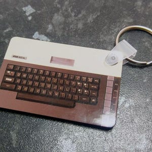 May include: A brown and white keychain featuring a miniature replica of an Atari 800XL computer. The keychain has a silver key ring attached.