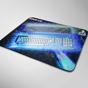 Puede incluir: Alfombrilla de ratón rectangular con un diseño retro de teclado y ratón de ordenador en azul y blanco. La alfombrilla tiene el logotipo de Atari y el texto "ATARI STE". El fondo presenta un patrón de placa de diamante.
