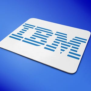 Op de afbeelding: Witte rechthoekige muismat met het blauwe IBM-logo. Het logo toont de letters "IBM" in een vetgedrukte, schreefloze letter, waarbij de letters worden gevormd door horizontale blauwe lijnen. De muismat ligt op een blauwe gradiënt achtergrond.