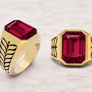 Puede incluir: Dos anillos dorados con grandes piedras rojas rectangulares. Uno tiene un diseño de hojas en el lateral, el otro en la parte superior e inferior. Los anillos están sobre un fondo blanco.