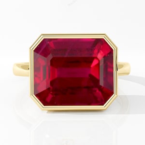 May include: A gold ring featuring a large, rectangular-cut ruby gemstone. The ruby is a vibrant red color and is set within a gold bezel. The ring has a simple, elegant design.