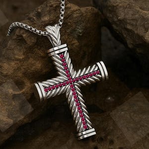May include: A silver cross pendant with a textured design and a chain. The cross features a row of red gemstones down the center and is set against a natural stone background. A religious jewelry item.