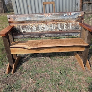 May include: A rustic wooden bench with a natural edge seat and armrests. The bench is constructed with dark wood legs and a back made from a vintage Chevrolet truck tailgate. The word "CHEVROLET" is visible on the back.