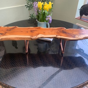 Puede incluir: Una bandeja de servir de madera natural larga con un borde vivo y patas de tubo de cobre. La bandeja está sobre una mesa de mimbre negra.