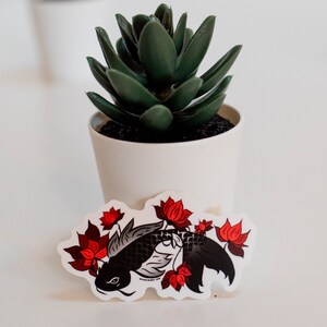 Puede incluir: Una pegatina con un dise&ntilde;o de carpa koi negra y flores de loto rojas. La pegatina tiene un borde blanco y el texto "MUNCHIES INK". Una peque&ntilde;a planta suculenta en maceta est&aacute; en el fondo.