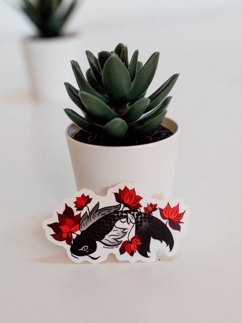 Puede incluir: Una pegatina con un dise&ntilde;o de carpa koi negra y flores de loto rojas. La pegatina tiene un borde blanco y el texto "MUNCHIES INK". Una peque&ntilde;a planta suculenta en maceta est&aacute; en el fondo.