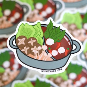 May include: A sticker of a hot pot with a variety of ingredients, including mushrooms, lettuce, and chili peppers. The hot pot is divided into two sections, one with a red broth and the other with a clear broth. The sticker is white with a gray pot and the text "MUNCHIES INK" at the bottom.