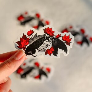 Puede incluir: Una pegatina con un dise&ntilde;o de pez koi negro con flores de loto rojas. El pez tiene escamas detalladas y est&aacute; rodeado de flores rojas vibrantes. La pegatina tiene un borde blanco y una apariencia ligeramente transparente.